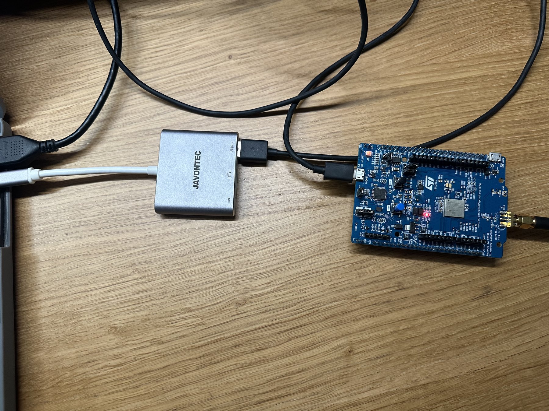 The STM32 B-L072Z-LRWAN1 Discovery kit on a wooden desk — a long blue development board with the square Murata radio module and an SMA antenna connector at one end — connected by a micro-USB lead through a small silver USB hub to a laptop off the left of the frame.
