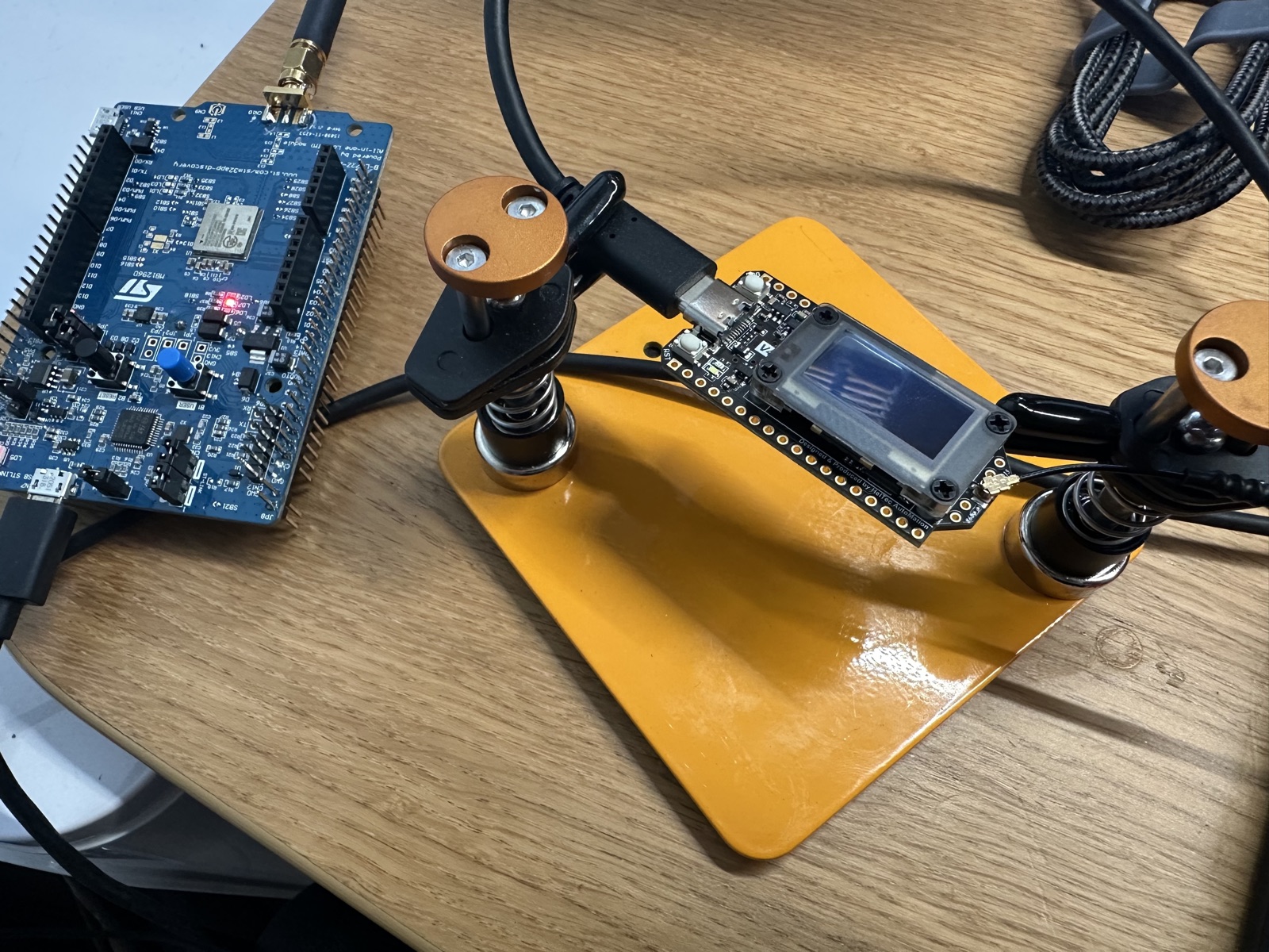 The blue STM32 B-L072Z-LRWAN1 Discovery kit wired by USB on the left, and the diymore LoRa V4, a small ESP32-S3 board with an OLED and a short stub antenna, clamped in helping-hands on a yellow work mat on the right, both on a wooden desk.